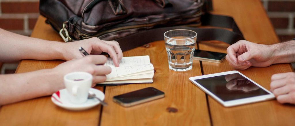 Twee mensen overleggen aan tafel met een notitieboekje, koffie en een tablet