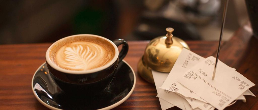 Een cappuccino met latte art naast een tafelbel en kassabonnetjes